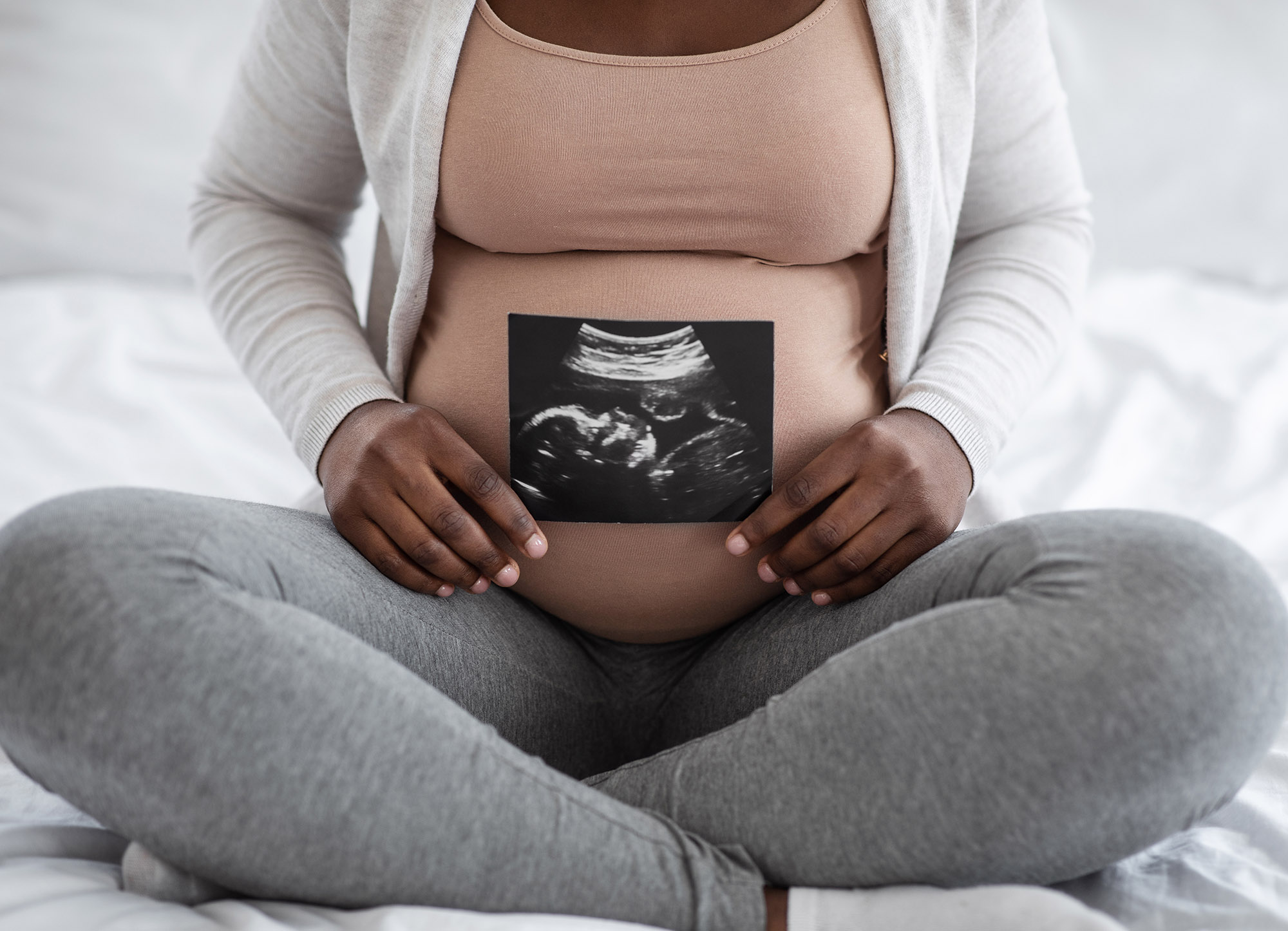 Photo: Unrecognizable Black pregnant person presenting their baby bump and baby sonography photo while sitting on bed at home.