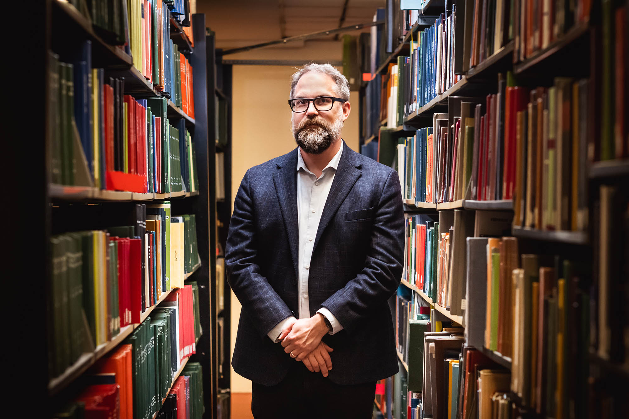 Photo: Mark Newton, a white man with dark and light gray hair, beard, and mustache and wearing glasses, a light blue collared shirt, and navy blue blazer and pants, poses with hands clasped in front of him between two large large library shelves filled with books.