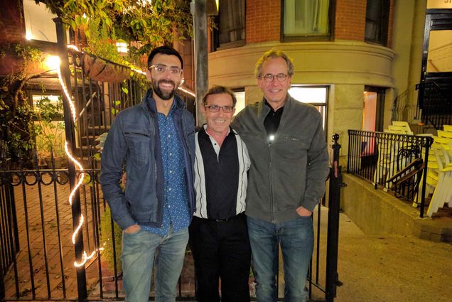 Photo: Three men stand to take a photo. It is night time and there fairy lights behind the one that stands on the left. From the left, a taller man with dark hair and a beard wears a denim button-up and a denim jacket. In the middle, a shorter man with dark hair and a black and white button-up, and lastly, on the right, a taller man with graying hair, a black t-shirt, and a gray sweater. They all wear glasses and are smiling for the photo.