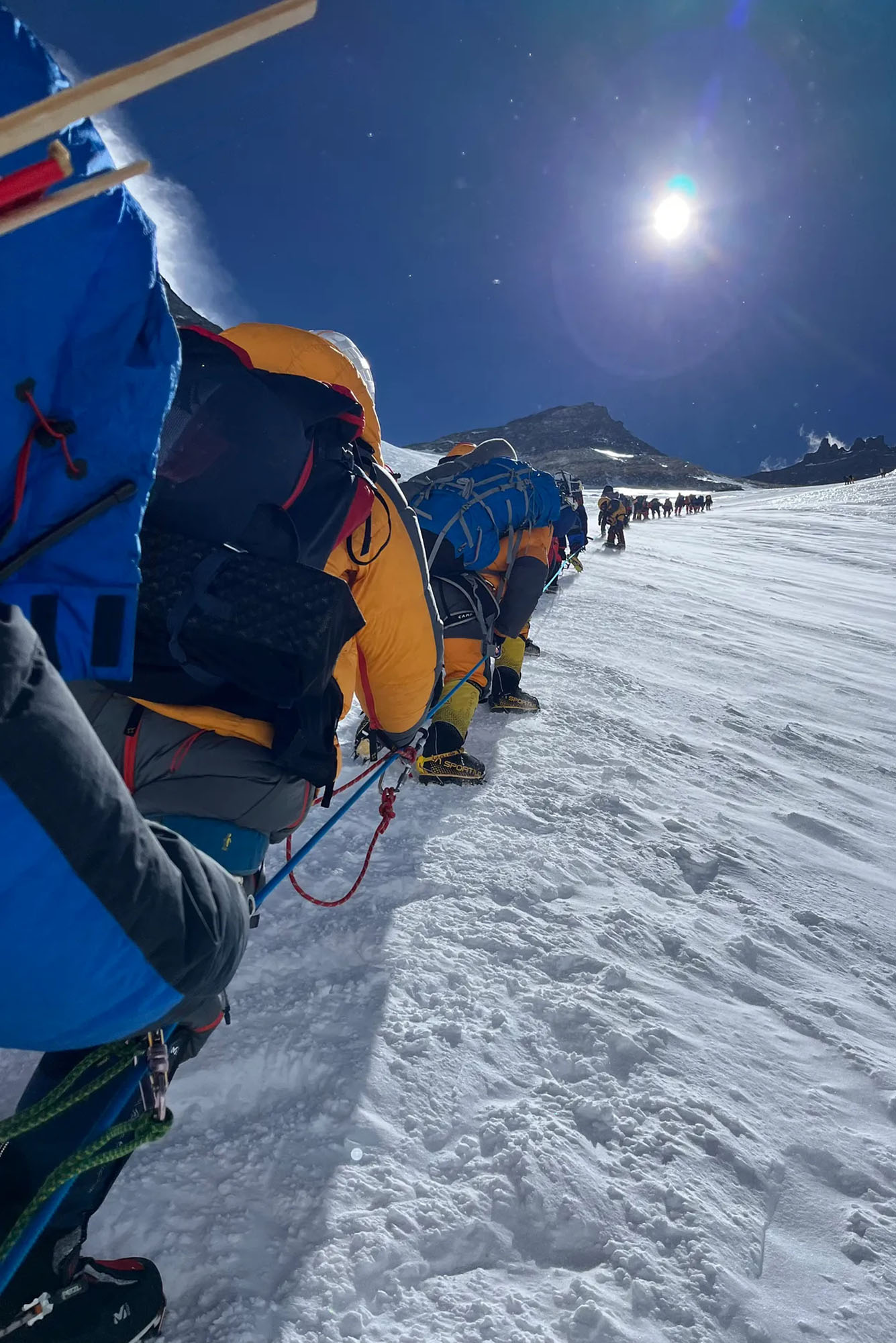 Photo: A vertical image of individuals climbing up a steep area on Mt. Everest. The climbers have various different gear on and the trail up snow-covered land. The sun shines bright above them.