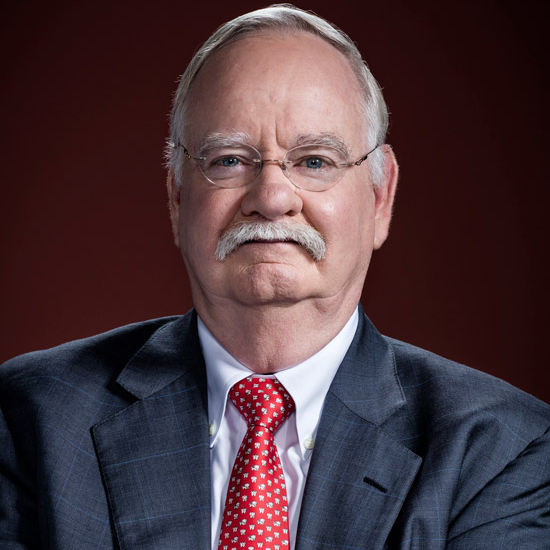 Photo: Headshot of Robert A. Brown, an older white man with silver grey hair and mustache and wearing glasses, a white collared shirt, red tie, and dark grey blazer. He smiles and poses in front of a burgundy red background.
