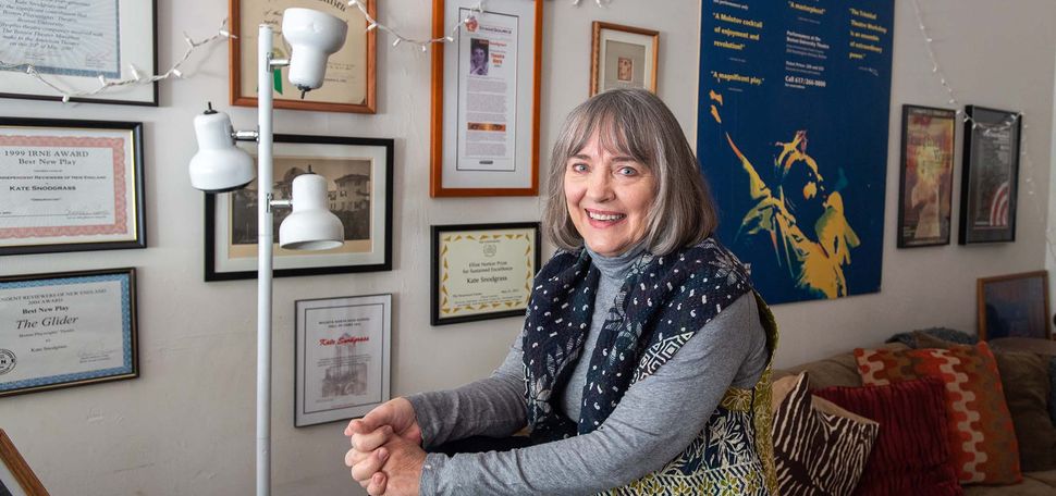Photo of Kate Snodgrass, a middle aged White woman with a gray bob, who wears a dark blue scarf with white pattern. She sits on the corner of a couch, presumably in her office, where framed awards and playbills hang on the wall behind her.