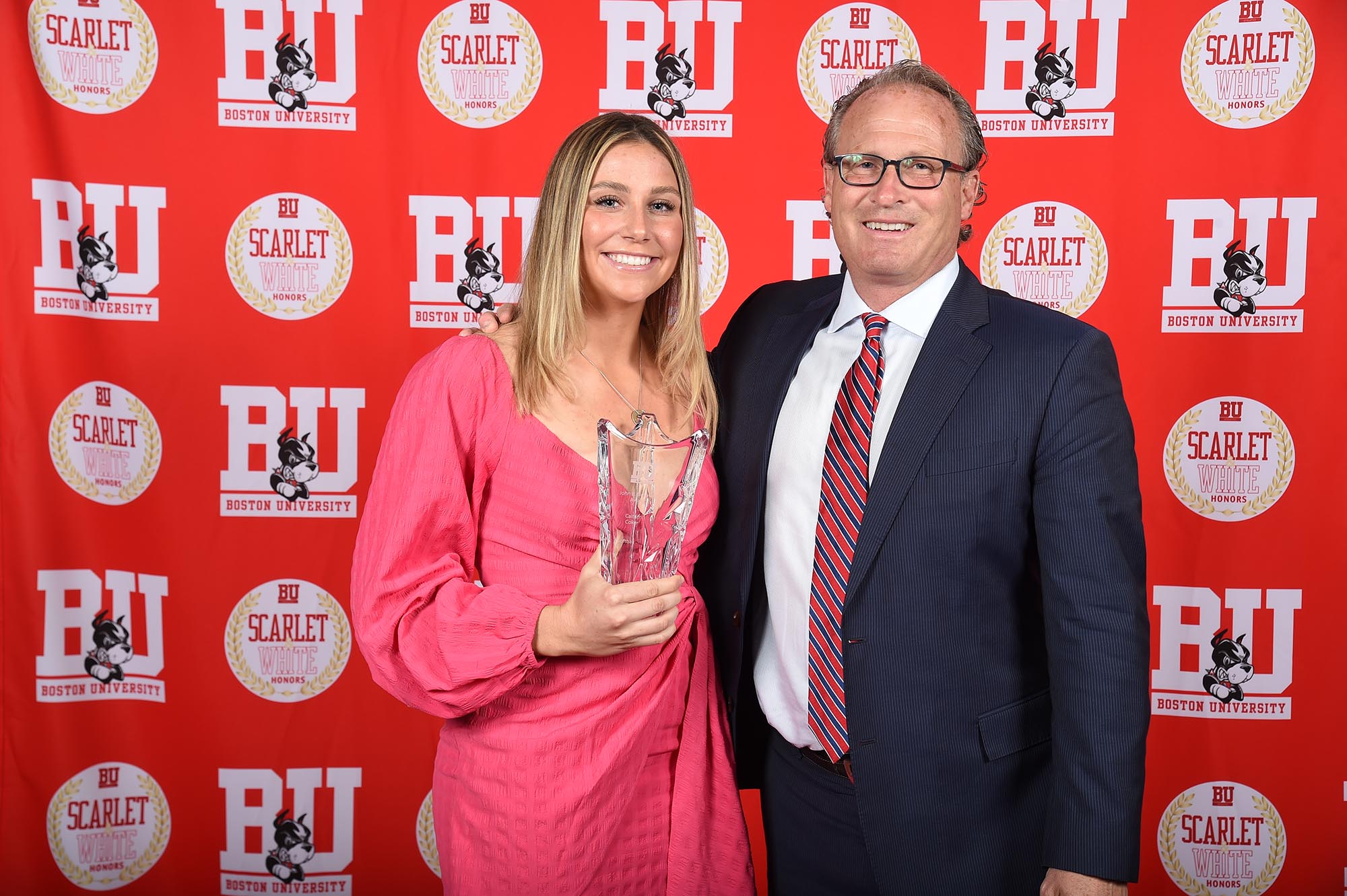Photo: Coker (left), a white woman wearing a pink dress and holding a glass trophy, stands with Drew Marrochello (right) a white man wearing glasses, a navy blue suit, white collared shirt, and red and blue striped tie, both smile and pose for a photo in front of a BU Athletics-themed backdrop.