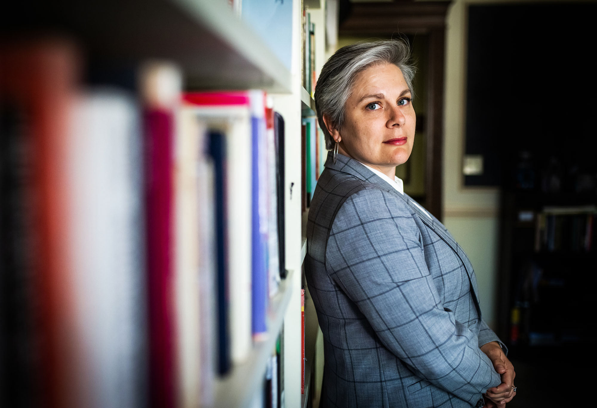 Photo: Cari Babitzke, a white woman with short silver-grey hair and wearing a blue-grey blazer and white collared shirt, leans her back against a bookcase and looks to the camera on her right.