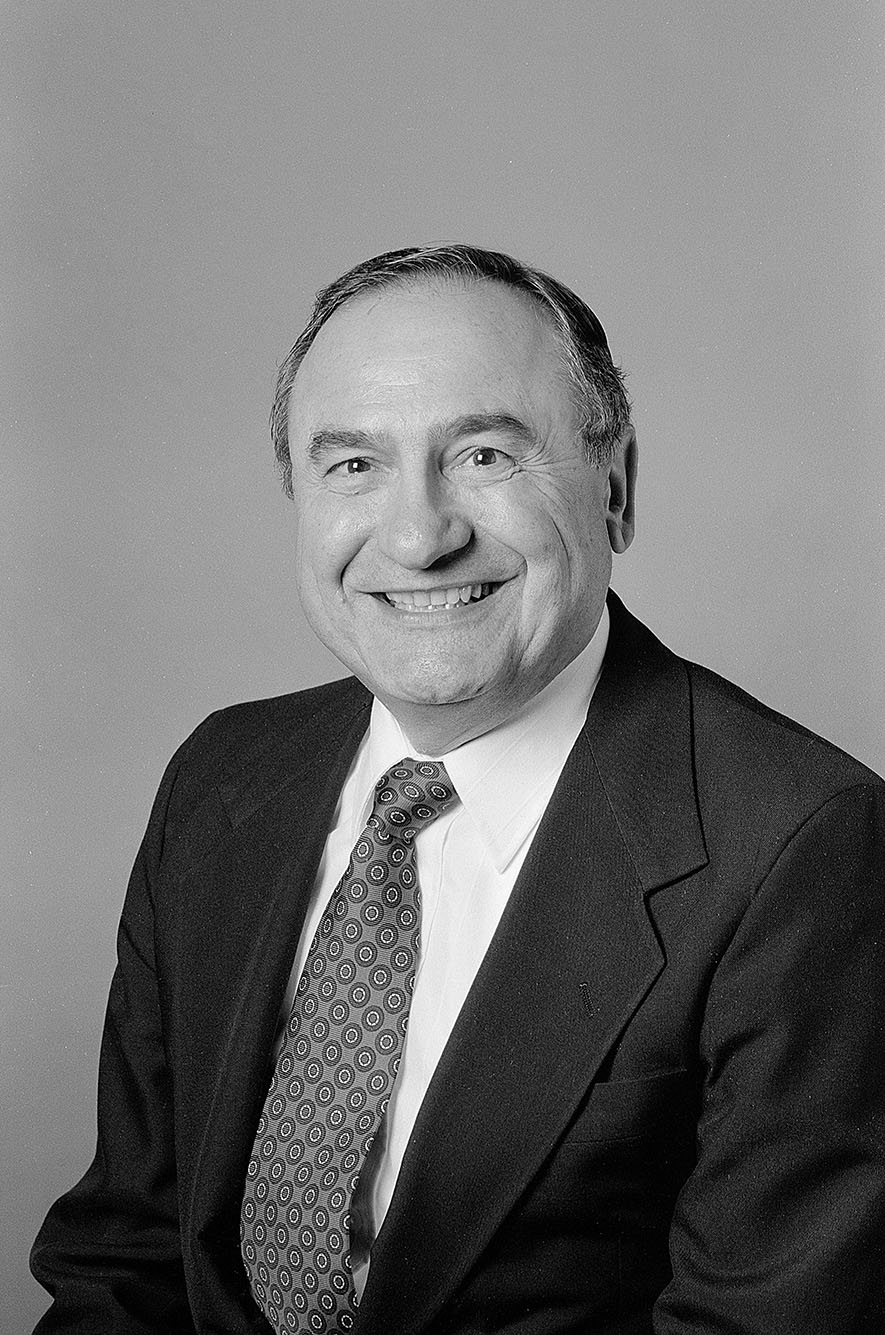 Photo: Black and white headshot of Christopher Barreca, a white man wearing a black suit, white collared shirt, and polka dot tie. He smiles and poses in front of a light-colored backdrop.