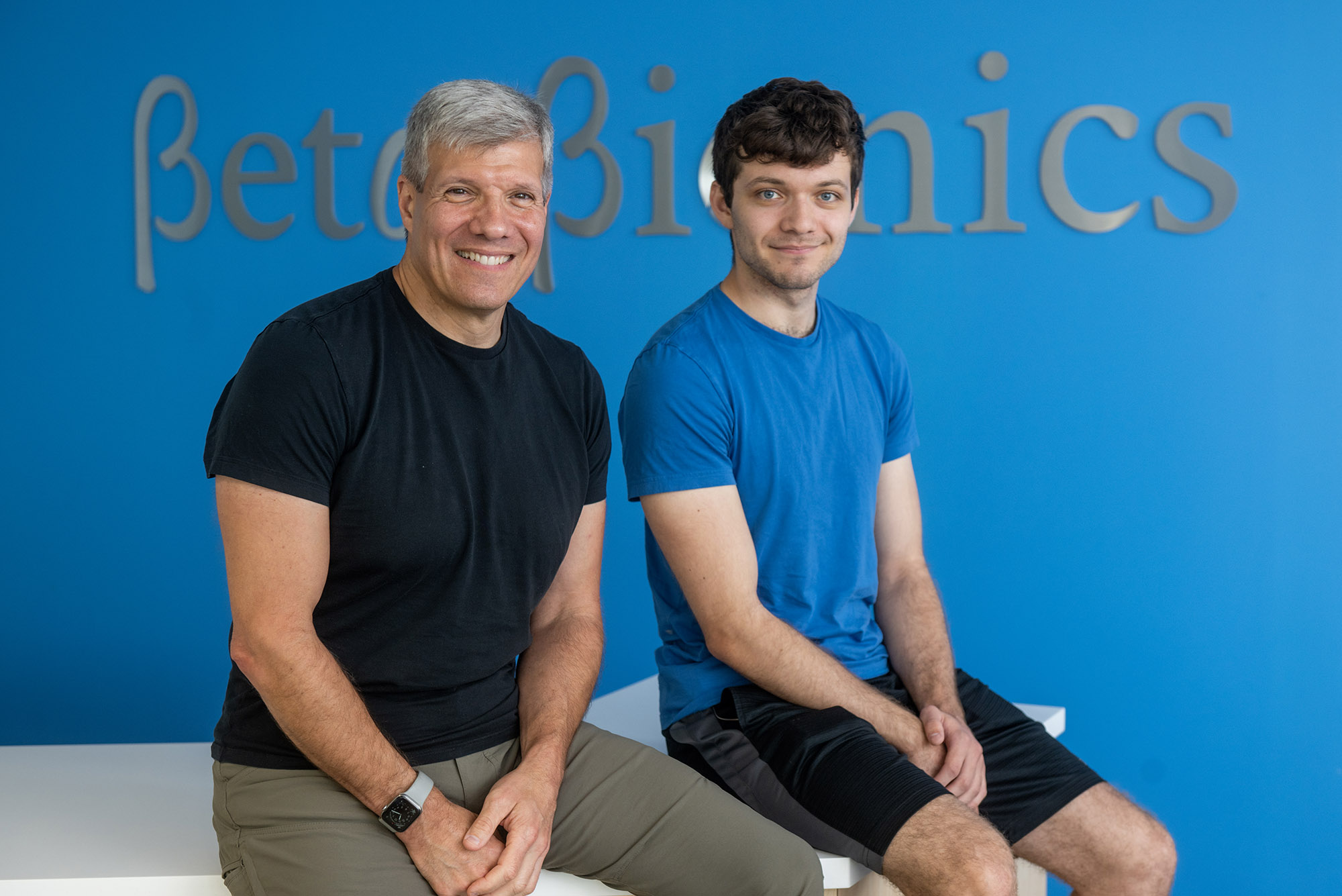 Photo: Beta Bionic’s Ed Damiano (left) with his son, David Damiano. A white man silver grey hair and wearing a black shirt and grey pants sits and poses next to a younger man with dark brown hair who is wearing a blue shirt and black pants. Both smile and pose in fornt of a blue wall. Silver letters on wall read "Beta Bionic".