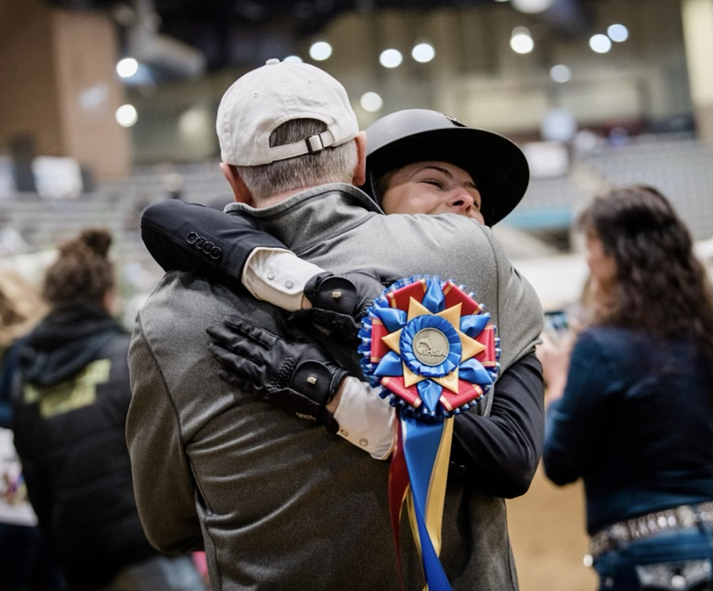 Photo: Elizabeth Nevins, a white young woman wearing black riding gear and helmet, is shown hugging a male figure wearing a brown leather jacket and white cap. She holds a large red and blue ribbon in her hand.