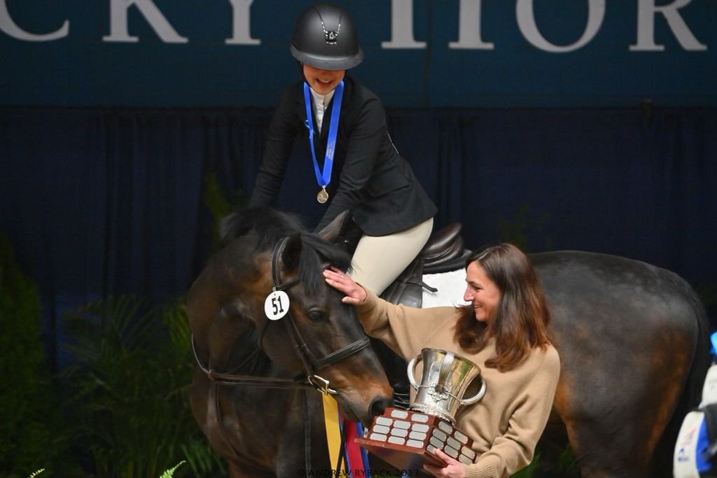 Photo: Elizabeth Nevins, a white young woman wearing black riding gear and helmet and gold medal, sits atop a dark brown horse as another white woman with brown hair and wearing a tan sweater, holds a large golden trophy and pets the horse.