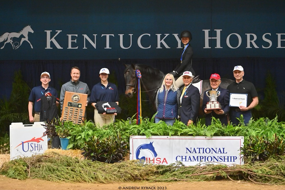 Photo: A group of people stand in a line holding various trophies, plaques, and awards. Behind them, Elizabeth Nevins, a white young woman wearing black riding gear and helmet, sits atop a dark brown horse. The sign in front of all of them reads "IHSA National Championship".