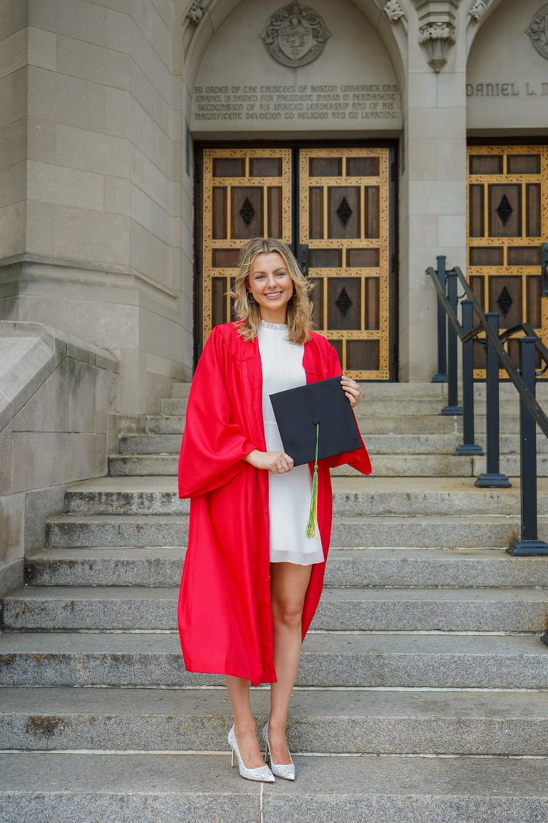 Photo: Elizabeth Nevins, a young white woman wearing a white dress and scarlet red graduation gown, smiles as she holds a black graduation cap and poses at the bottom of stone stairs that lead up to an intricate wooden door.