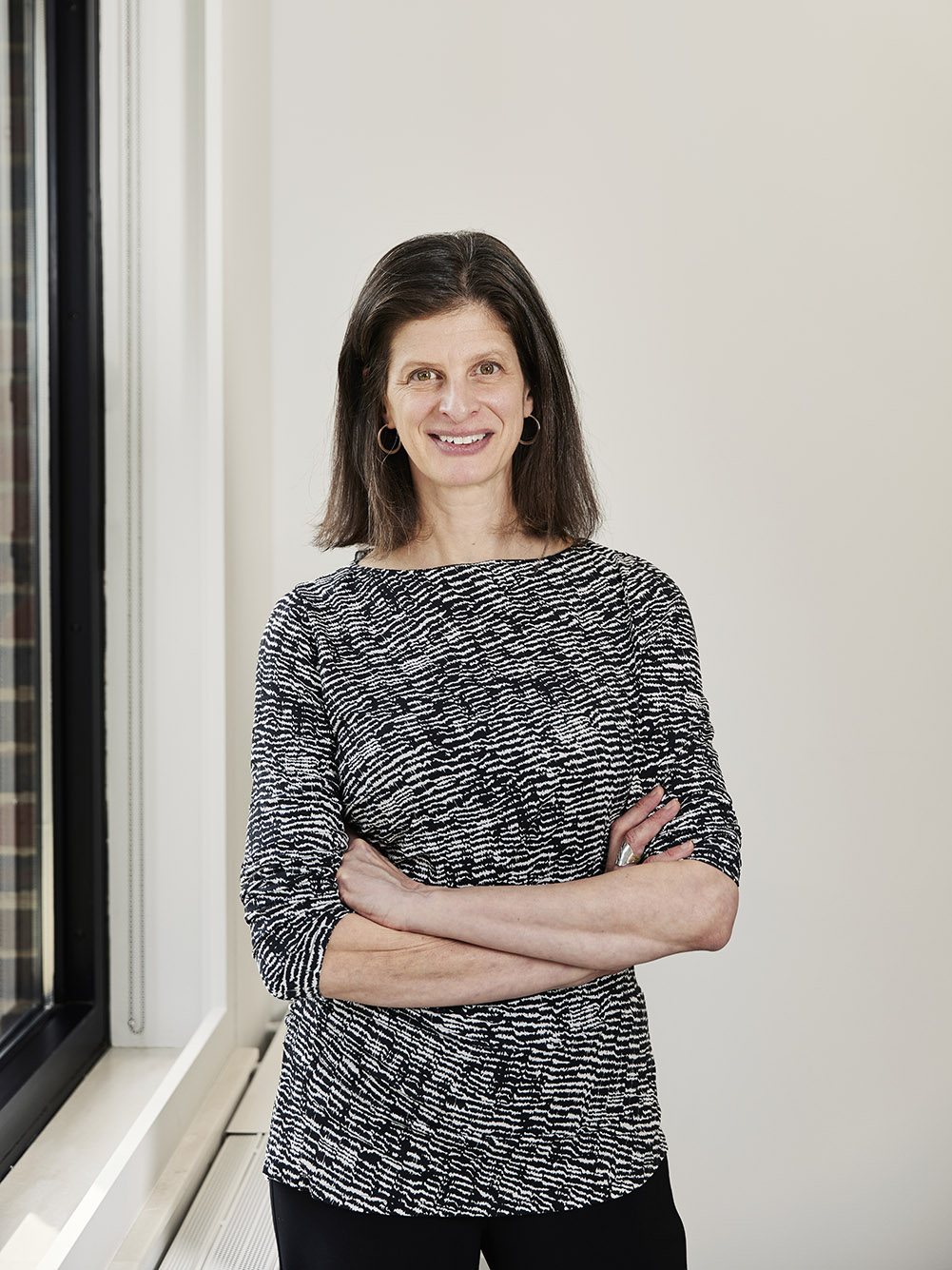Photo: Dr. Emily Rothman, a white woman with shoulder-length brown hair and wearing a black and white half-sleeved dress, stands and poses with arms crossed. She smiles as she stands next to a large window in a brightly lit space.