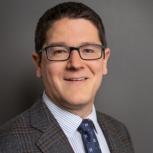 Photo: Headshot of Garrett Johnson, a white man wearing glasses, a collared shirt, dark blue tie, and dark grey blazer. He smiles in front of a grey backdrop.
