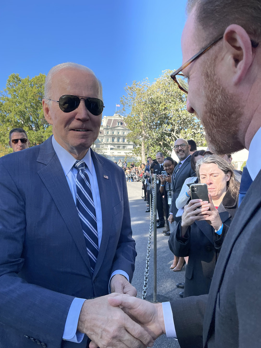Photo: Goodman (right) shaked hands with President Biden. Goodman stands to Biden's right on the other side of a small, metal separation. Biden smiles and wears dark black sunglasses as he shakes Goodman's hand.