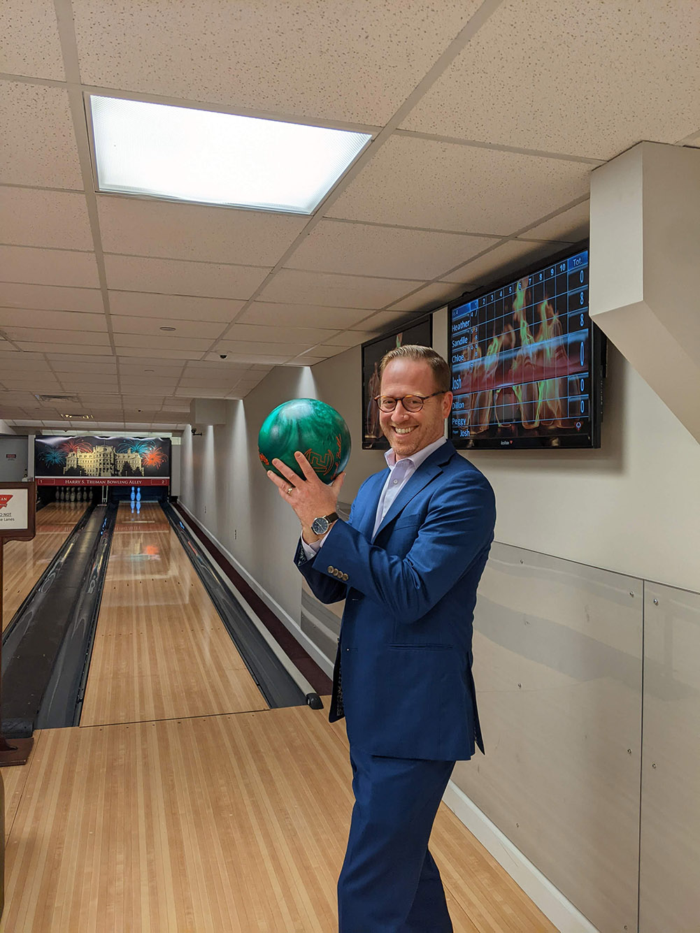 Photo: Goodman, a white man wearing a blue suit and glasses, holds up a green bowling ball and poses in front of a bowling lane.