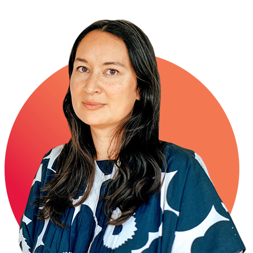 Image: : Michelle Min Sterling, a tan woman with long black hair and wearing a dark and light blue blouse, smiles and poses for a photo. Behind her is a gradient red and orange circle.