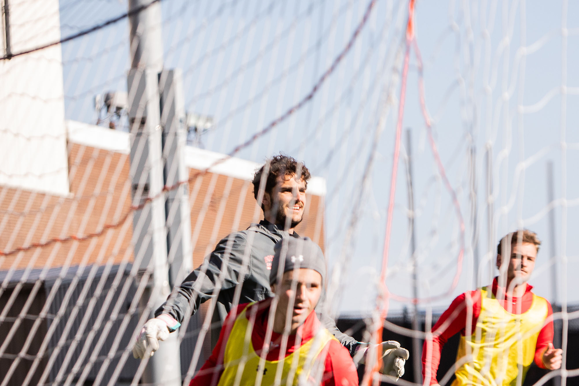 Photo: Action shot of Francesco Montali as he stands and raises his hand in a soccer goal. A young white man with curly brown hair and wearing a grey jacket and black short and spandex, jumps up as two young men wearing red jackets and yellow vest run ahead of him. Photo is shot behind the goal net.