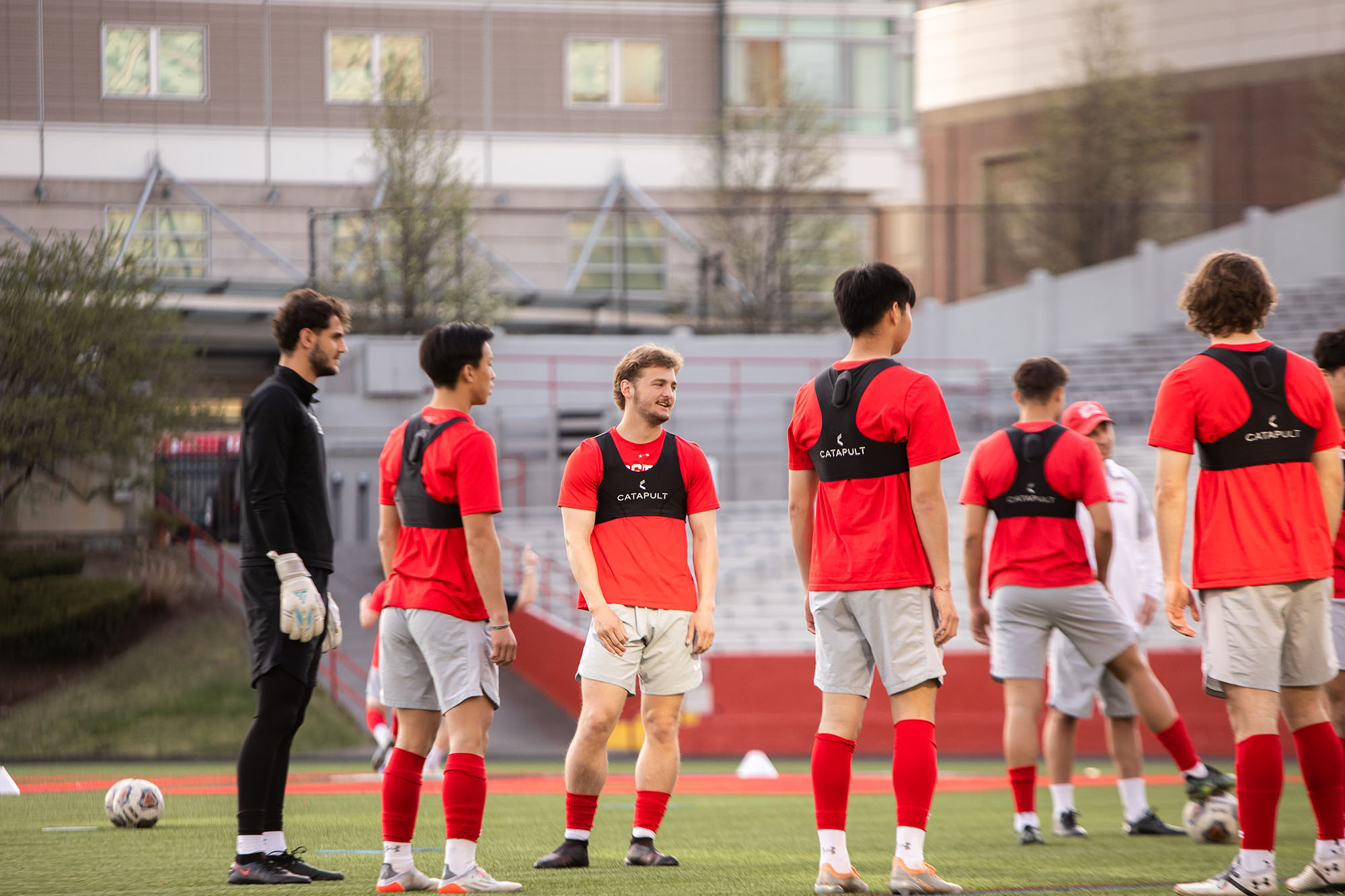 Photo: Francesco Montali stands with teammates amidst a soccer practice. A young white man with curly brown hair and wearing a black jacket, shorts, spandex, and white gloves, stands with teammates wearing red shirts and grey shorts.