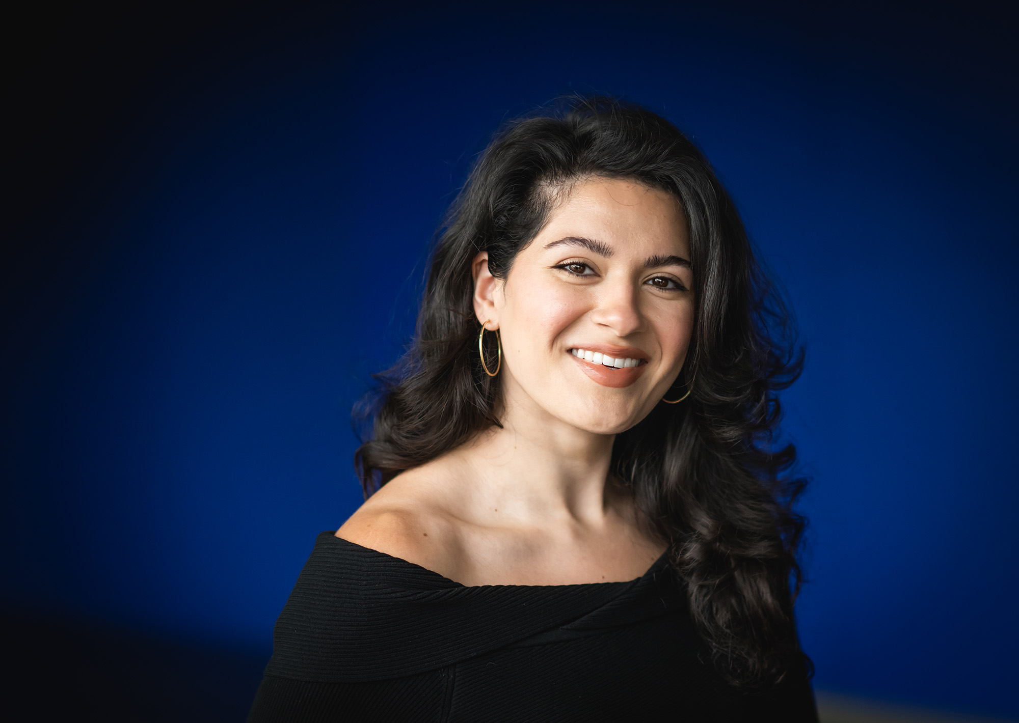 Photo: Nikta Khani, a young woman with long, curled black hair and wearing an off the shoulder black long sleeved top, smiles and poses in front of a dark blue backdrop.