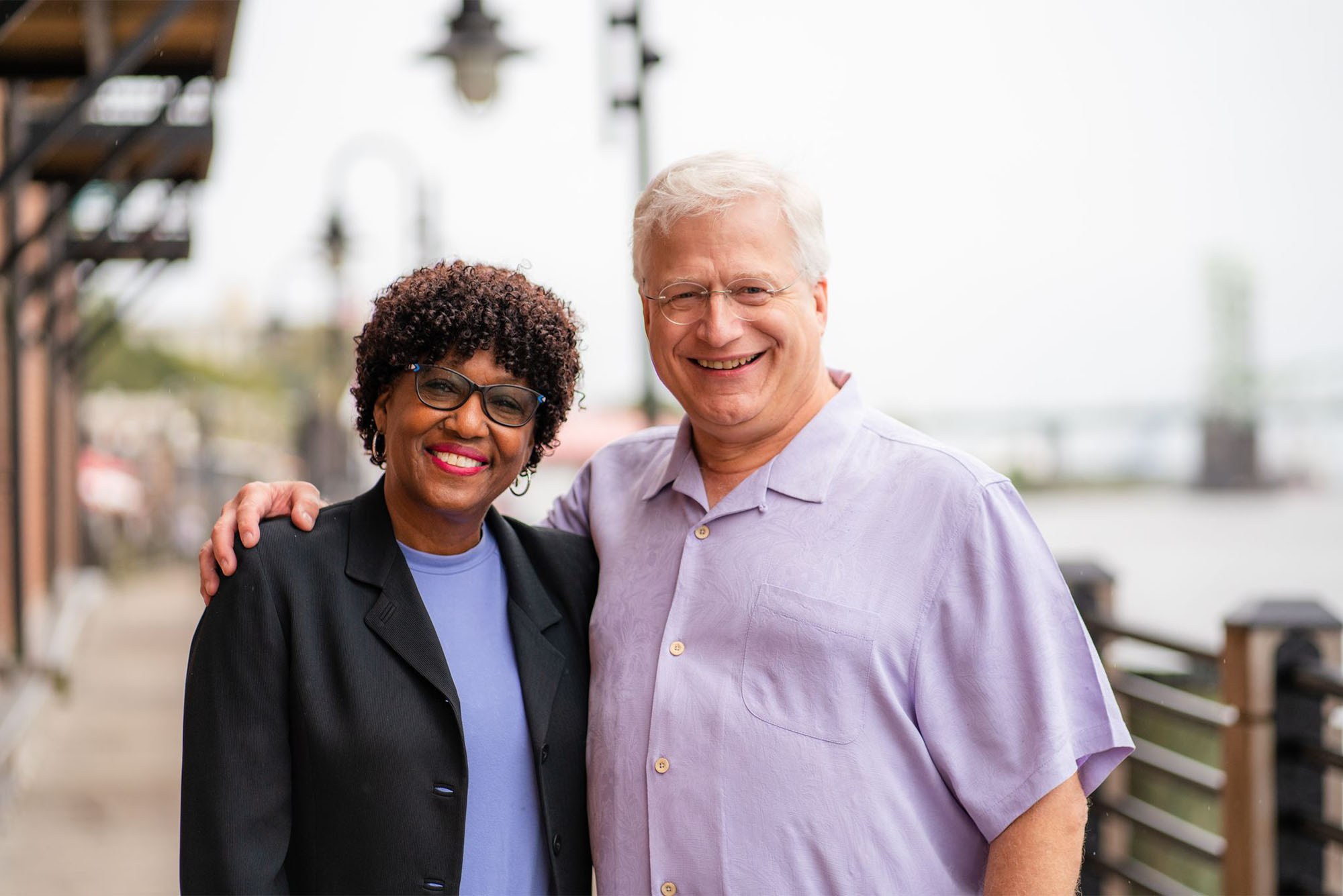 Photo: A black woman and white man stand side by side with smiles on their faces in front of a body of water on an overcast day