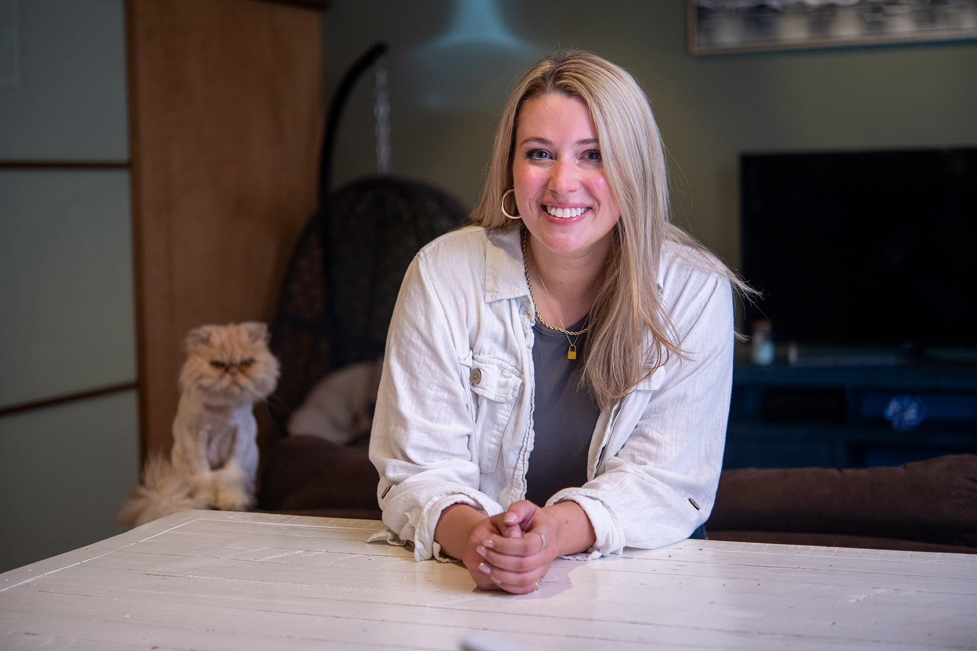 Photo: Stacey Cunnington, a white woman with long ash blonde hair and wearing a dark grey shirt and white jacket, sits and poses with hands clasped on the table in front of her. She was photographed in her Boston home. Behind her is one of her cats, a scruffy looking cat with a grumpy face.