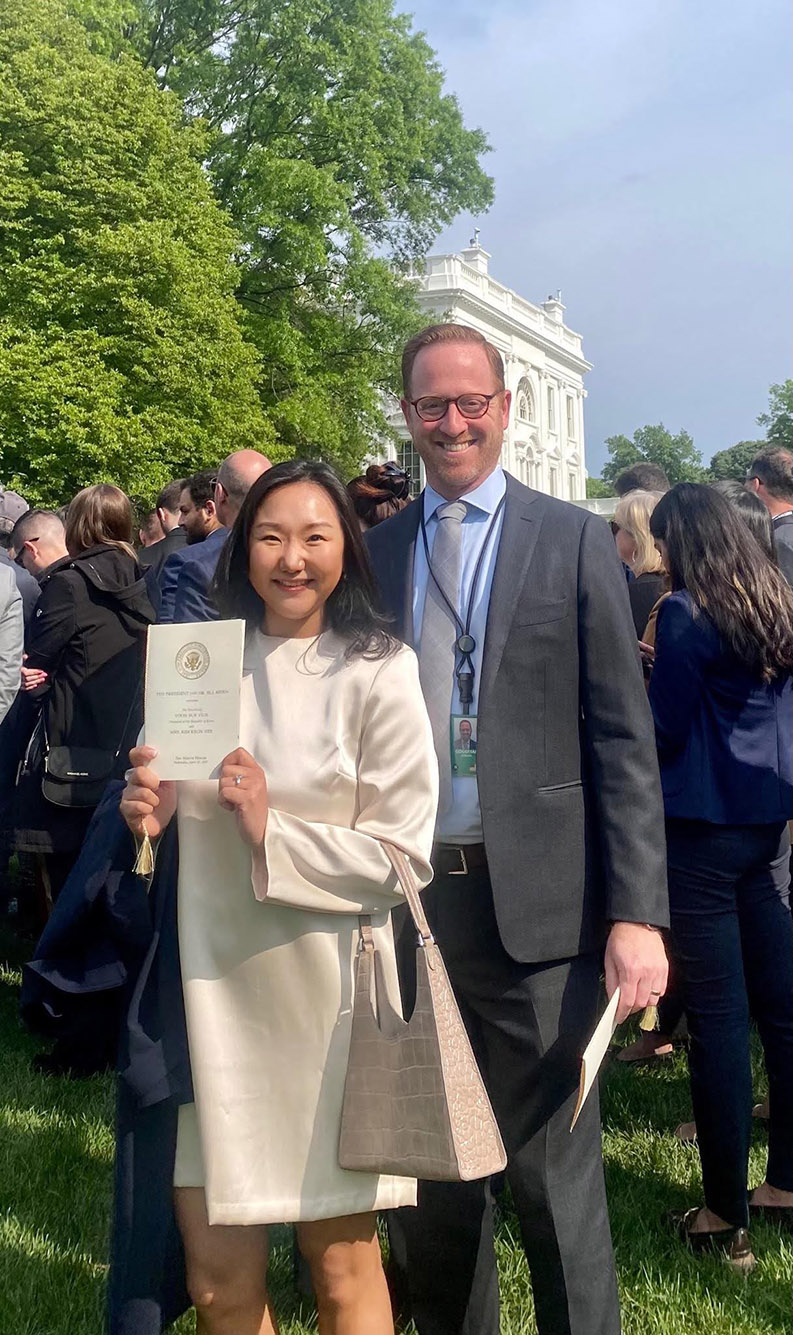 Photo: Goodman (right) poses with Yunee Yoon outside the White house. A white man (right) wearing glasses, a light blue collared shirt, and grey suit smiles and poses next to a young Asian woman holding up a white house pamphlet and wearing a long, cream-colored jacket.