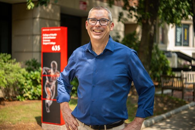 Photo: A white man in a blue button down smiles for the camera with his black glasses. He is outside and his hands are on his hips.