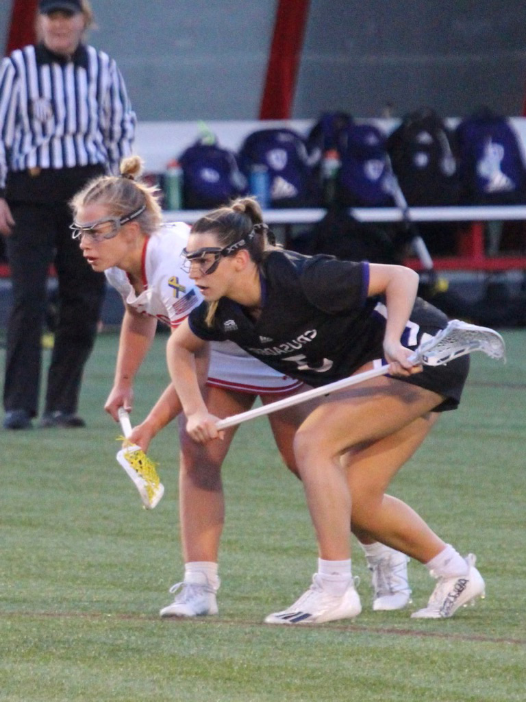 Photo: Action shot of Ellen Monahan as she plays lacrosse. A young white woman wearing a BU lacrosse uniform and goggles bends over and throws elbows with an opposing athlete wearing a black uniform. A referee can be seen on the field in the blurred background.
