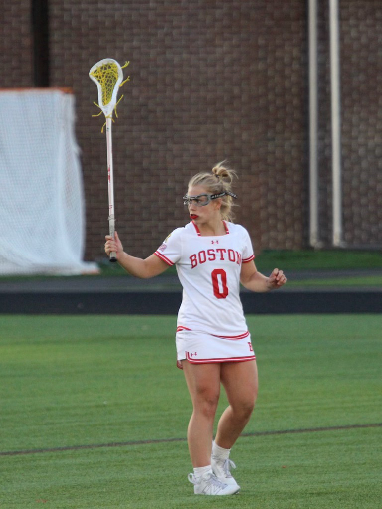 Photo: Action shot of Ellen Monahan as she plays lacrosse. A young white woman wearing a BU white and red lacrosse uniform and goggles looks to the left as she raises her lacrosse stick in her left hand.