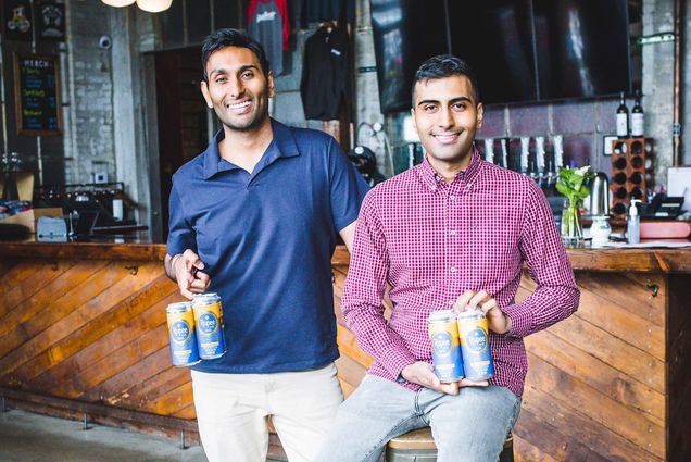 Photo of Sumit Sharma (right, in red) and his brother Vanit Sharma (at left, in blue) posing with four packs of Rupee beer at Dorchester Brewing Company. The brothers are Indian American, have dark hair, and smile as they pose. Sumit sits on a metal stool and Vanit stands. A wooden bar and taps are seen behind them.