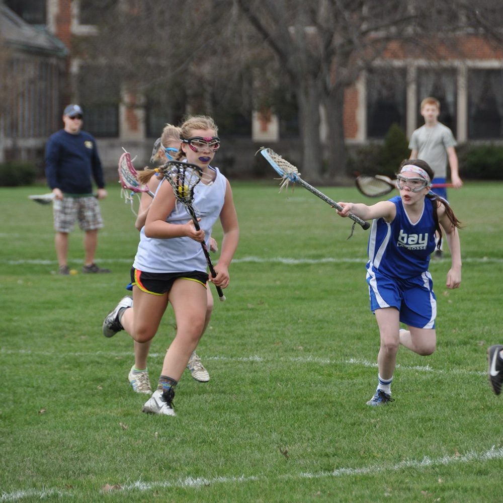 Photo: Action shot of young Ellen Monahan as she plays lacrosse. A young white woman wearing a white jersey, black shorts, goggles, and mouthguard runs down the field with a lacrosse stick holding a yellow ball. Another player in blue reacher her stick out to try and stop her.