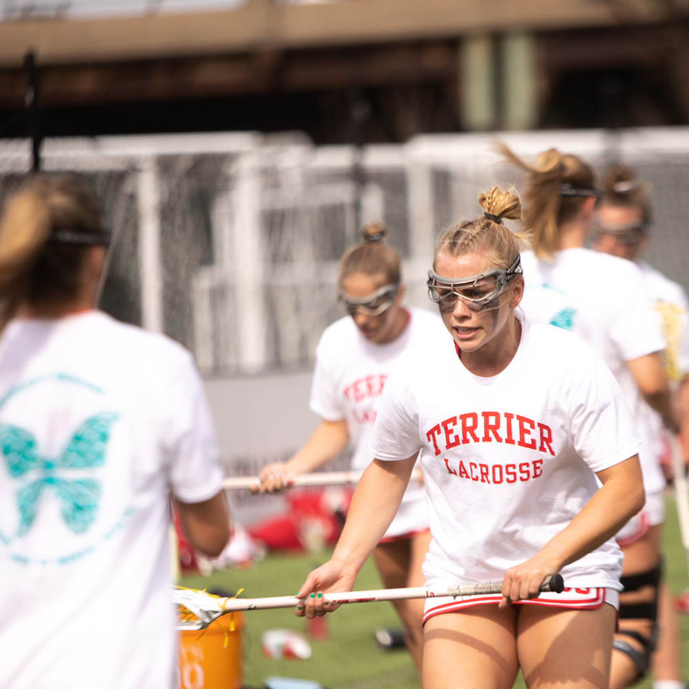 Photo: Action shot of Ellen Monahan as she plays lacrosse. A young white woman wearing a BU white and red lacrosse uniform and goggles looks forward as she practices catching balls with her lacrosse stick. Another athlete stands in the foreground throwing balls for her to catch.