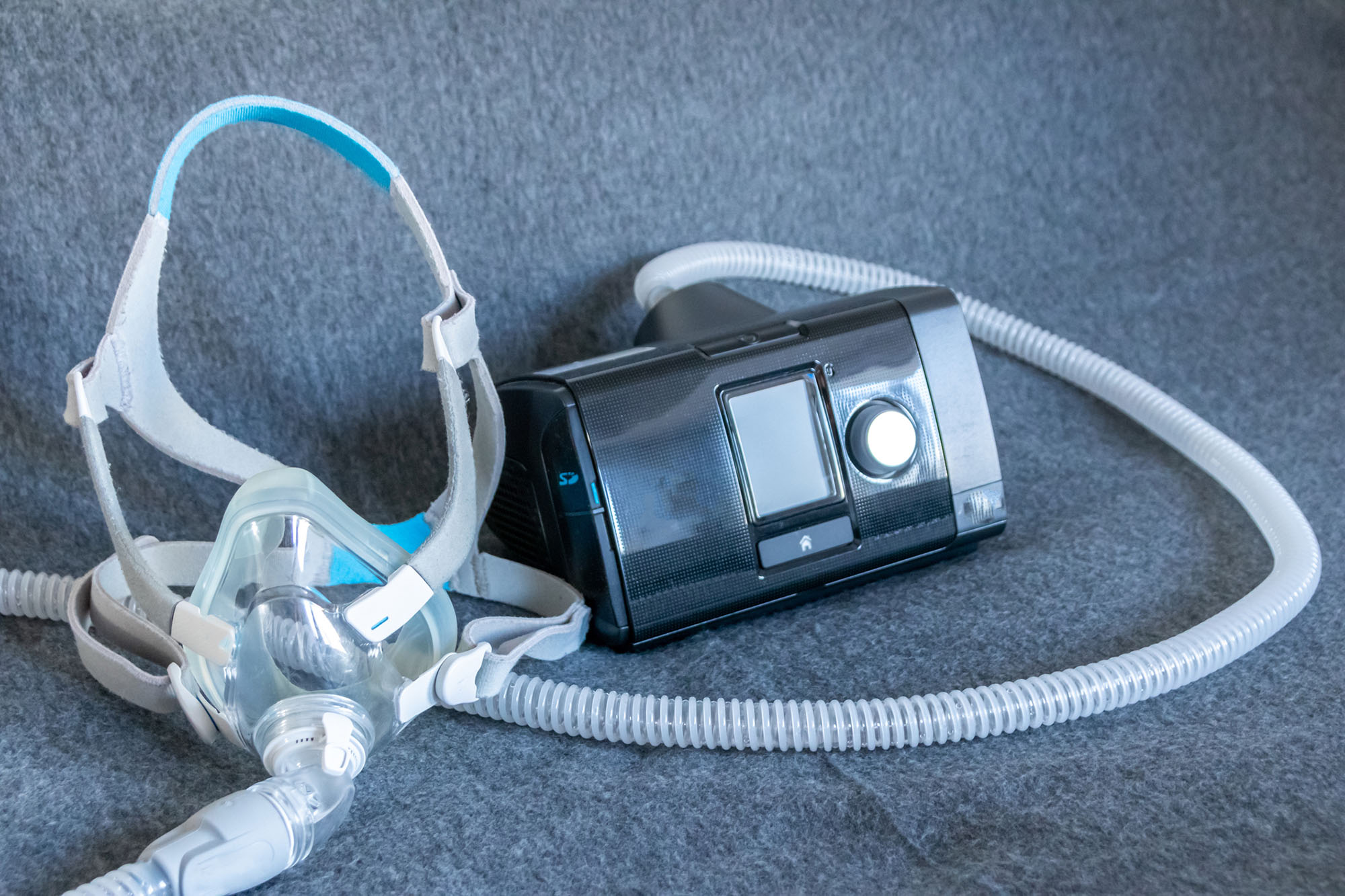 Photo: CPAP machine with a full face mask attached is shown on a grey background. The small, black machine has a simple interface with a hose attached the mask.