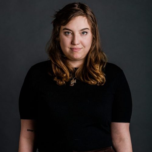 Photo: Headshot of Sophie Yarin. A white woman with wavy brown hair and wearing a black dress and gold necklace, smiles and poses in front of a dark grey backdrop.