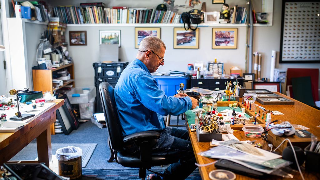 Photo: Scott Biron makes a fly at his New London, NH home. An older white man wearing glasses, a long-sleeved blue collared shirt, and jeans sits at a crowded desk and works on a fly. Fly tying materials and paraphernalia can be seen around the office.