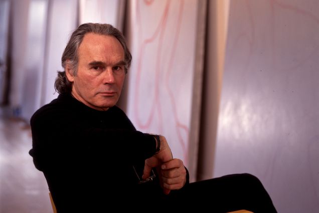 Photo: Portrait of Brice Marden, a white man with grey-brown hair and wearing black long sleeved shirt and pants, sits on a chair and looks over his shoulder at the camera. He sits in front of a wall full of large canvases featuring his paintings.