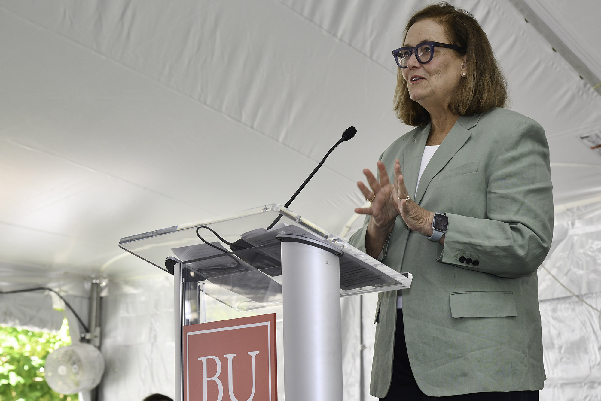 Photo: A white woman in a sage blazer talks at a podium with the letters BU in bright white and bold on the front. She has short brown hair and wears black framed glasses.