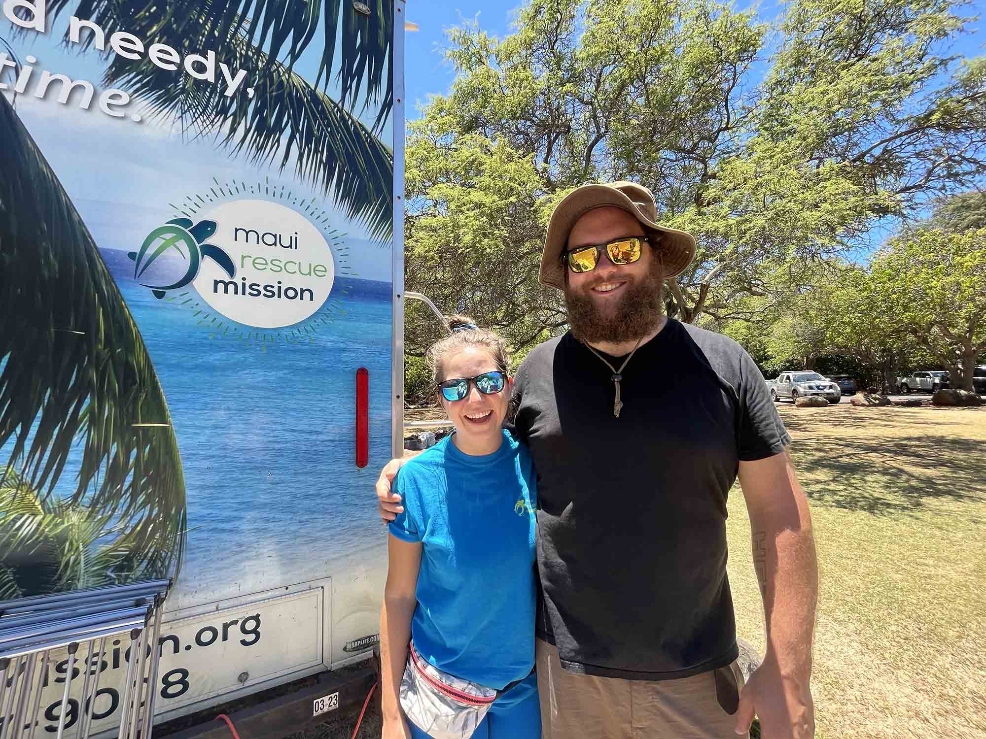 Photo: Lauren Wittel-Henrie and Ben Wittel-Henrie by the Maui Rescue Mission trailer. A white woman (left) wearing blue sunglasses, shirt, and shorts, smiles and gives a side hug to a white man wearing orange sunglasses, tan bucket hat, black shirt, and khaki shorts. They stand in front of a small trailer that reads "Maui Rescue Mission".