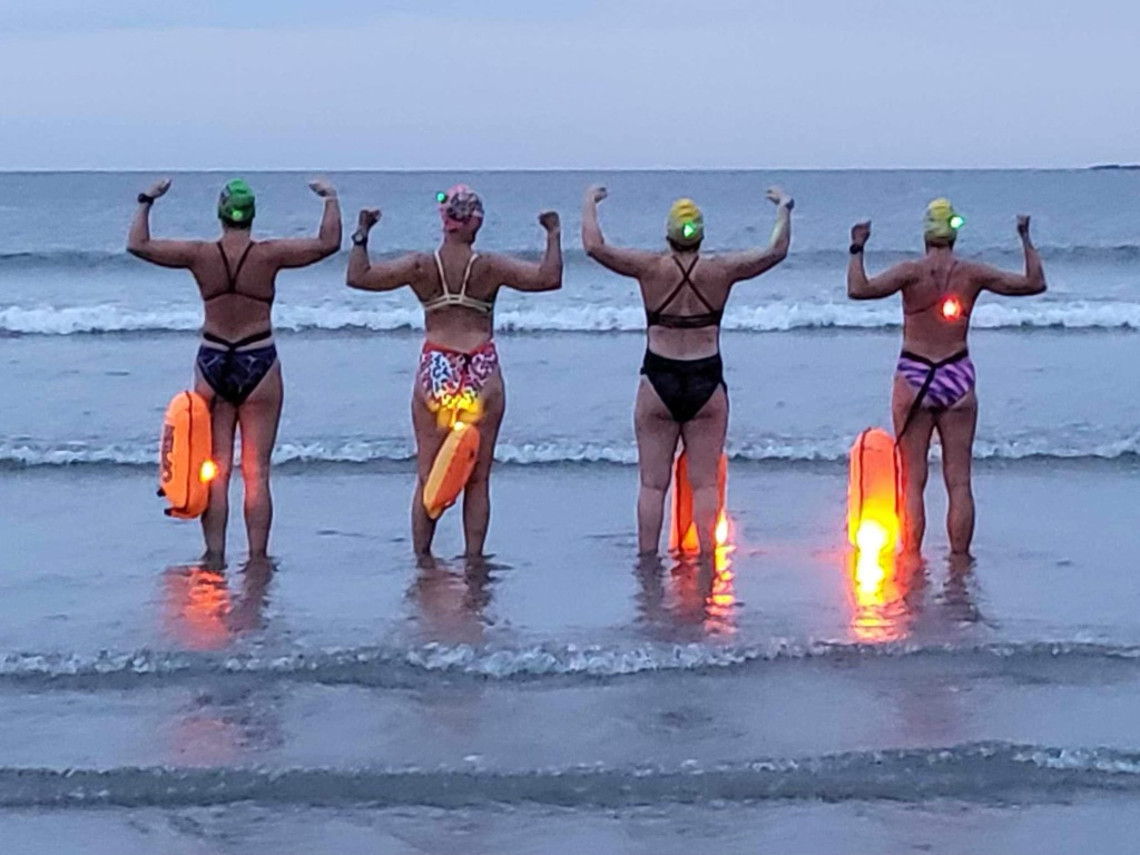 Photo: 4 women in swimsuits and swim caps walk with holding each other's hands with arms raised, walk on a shore towards an ocean at dusk. Small lanterns at their feet glow orange.