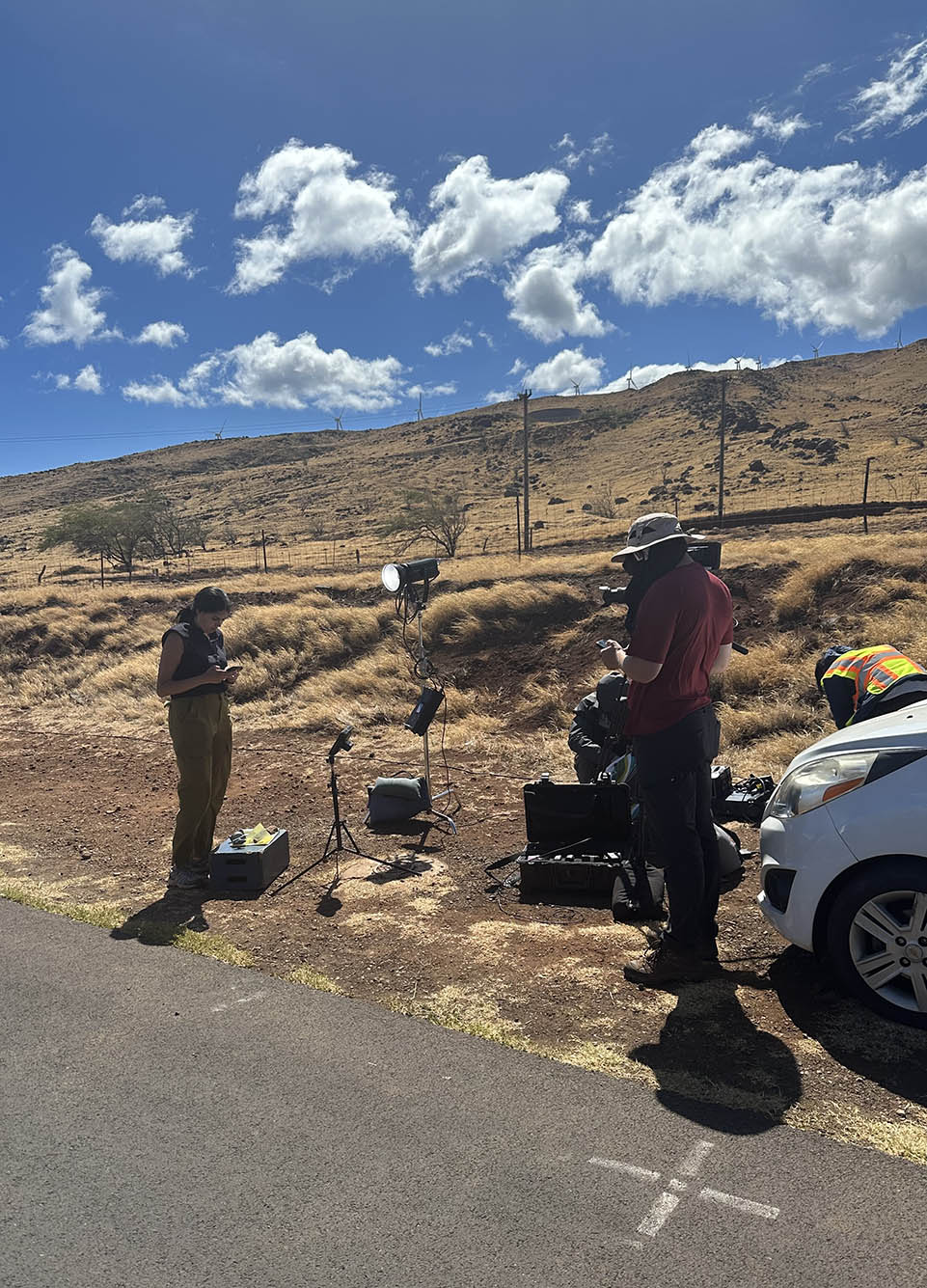 Photo: Melissa Adan of ABC News is shown reporting live from a roadside in Maui. A tan woman with black hair tied back and wearing a navy blue sleeveless blouse and khaki pants looks to her cellphone as a man wearing a large bucket hat, red shirt, and jeans holds a large TV camera in front of her. they stand on a dry, dirt road as fluffy clouds litter the blue sky.