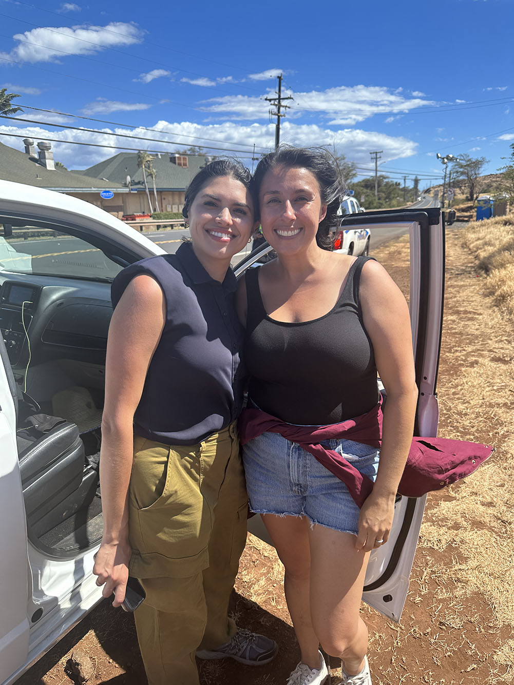Photo: Adan (left) and Hooper met up for the first time in years on that Maui roadside. A tan woman with black hair tied back and wearing a navy blue sleeveless blouse and khaki pants smiles and hugs with one arm another tan woman to her right. The other woman wears a black tank top and jean shorts. They stand by the side of an car on a dry, dirt road.