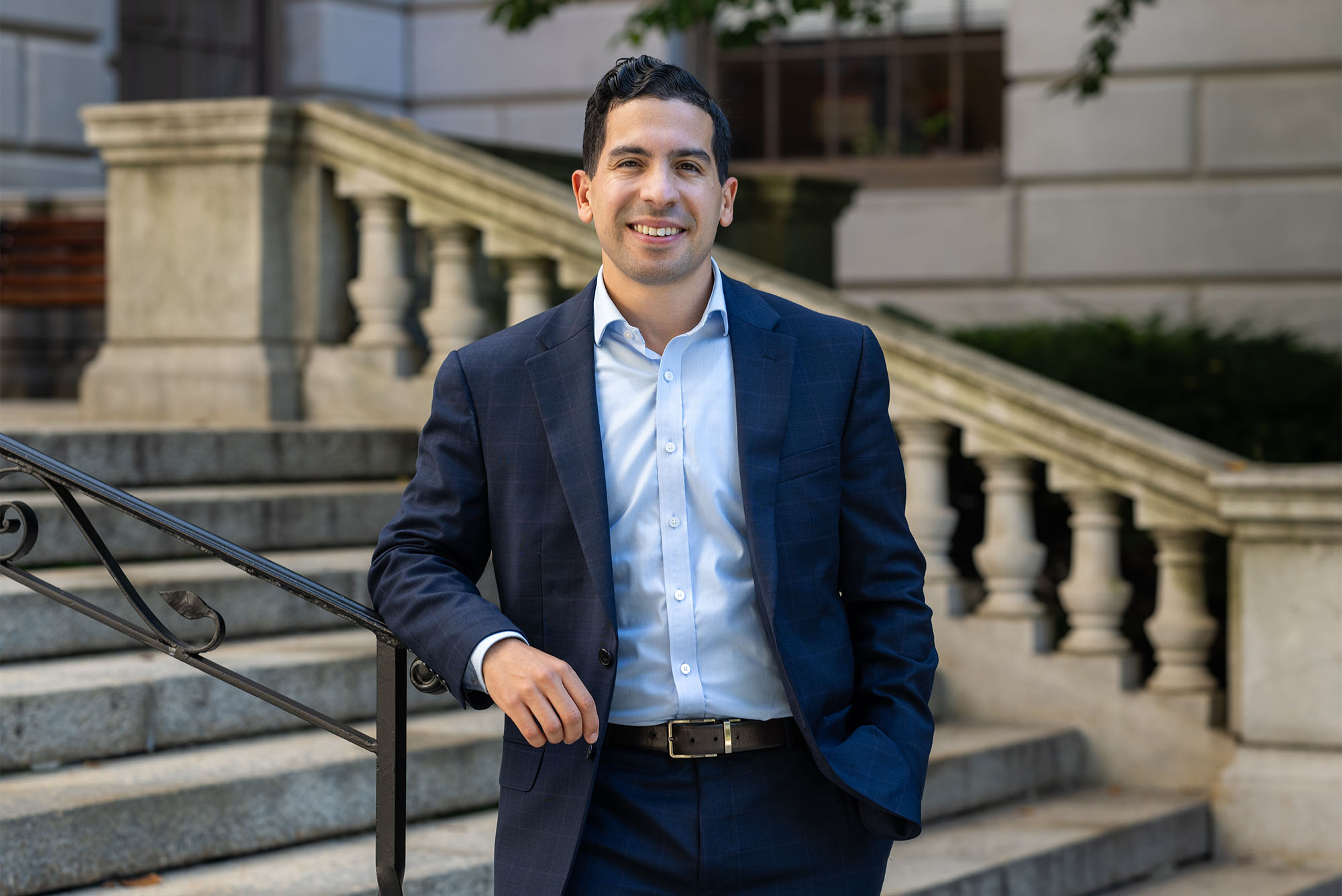 Photo: A man in a navy suit with short dark hair smiles on the steps of the Massachusetts State House, a large ornate government building.