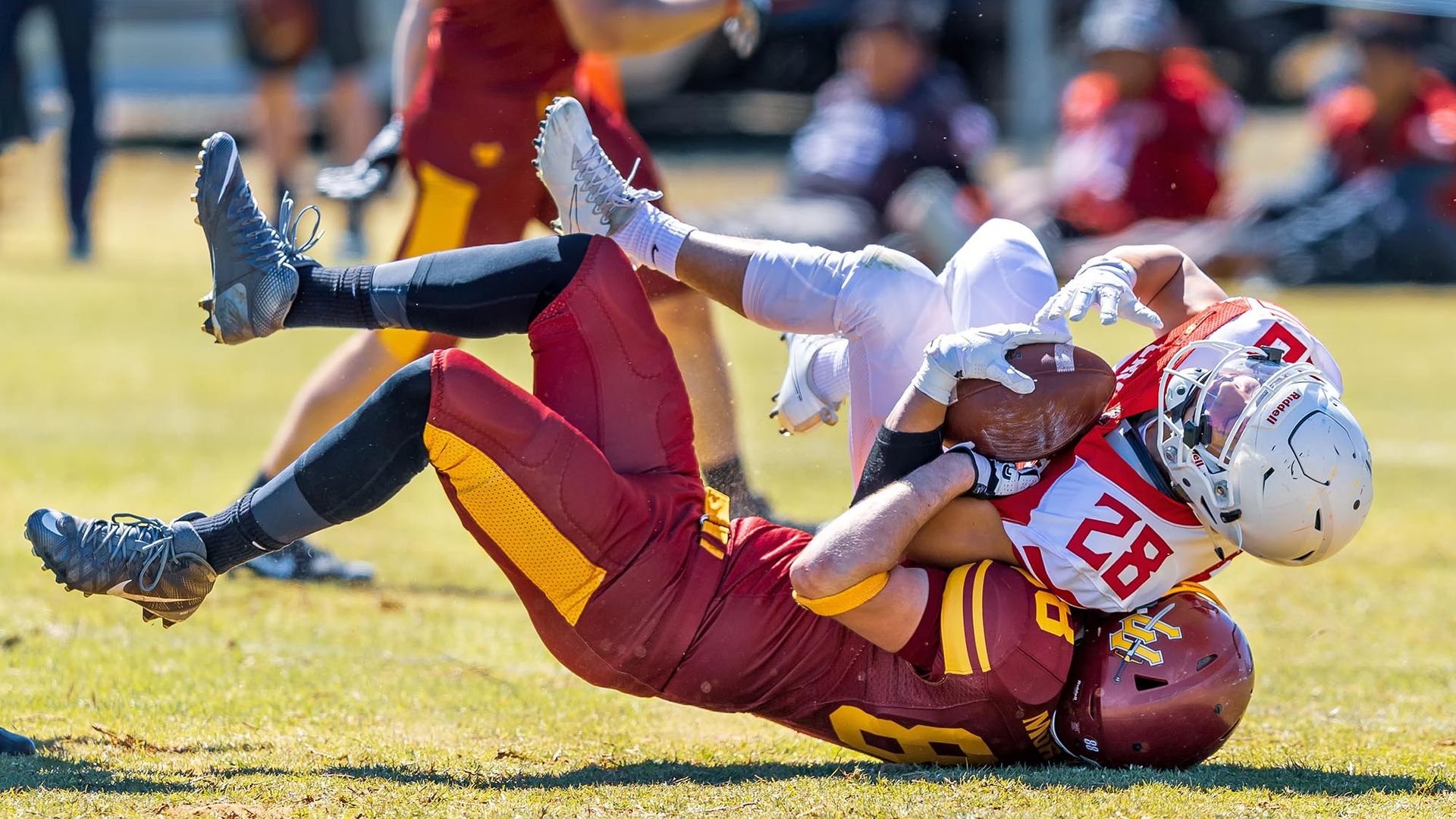 Photo: A person wearing a burgundy and yellow tackle football uniform and helmet is shown tackling a player in a white and red uniform and helmet tot he ground. The burgundy athlete has their head impacted to the ground as the white player falls on top of them.