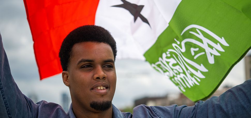 Photo: Zakariya Hussein, a dark-skinned Somalian young man wearing a black shirt and blue collared lone-sleeved shirt smiles and holds the Somaliland flag above his head. The flag blows behind him, creating a striking portrait.
