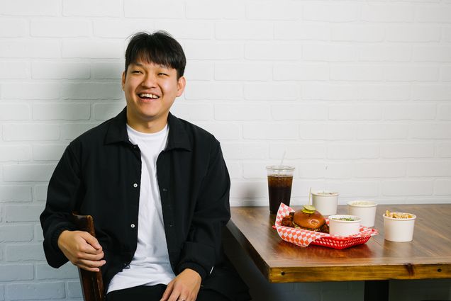Photo: An Asian man with short black hair smiles as he sits in a chair next to a standard order at his chicken shop.