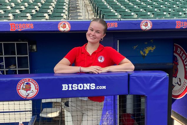 Photo: A young white woman with a red collared shirt and khakis leans on a blue fence in front of stadium seats. She smiles for the camera.