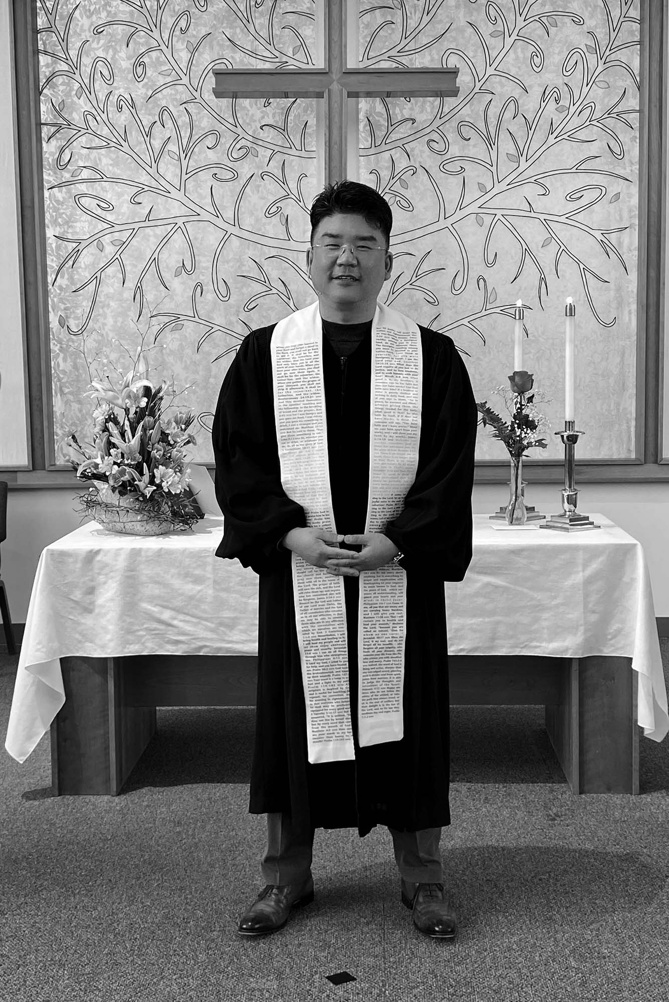 Photo: A Korean man, Jinwoo Chun, clasps his hands in front as he stands to pose for the photo. He wears his religious robes in front of a table with a white tablecloth in a cross on the background.