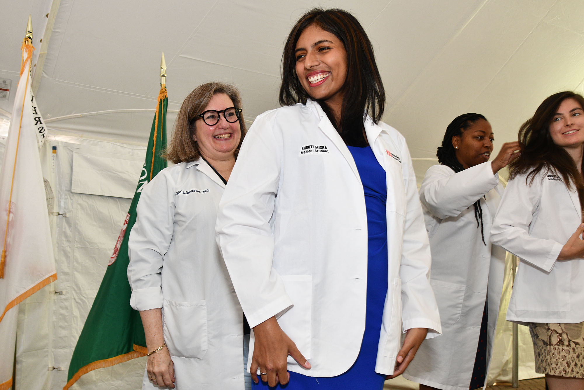 Photo: A woman with a blue dress and short, dark hair wears a white lab coat in front of a woman with short, light brown hair at a white coat ceremony. To the right of the two individuals in the center are two other individuals in the exact position, both smiling as the one behind helps the one in front don her coat. They are standing on a covered stage.