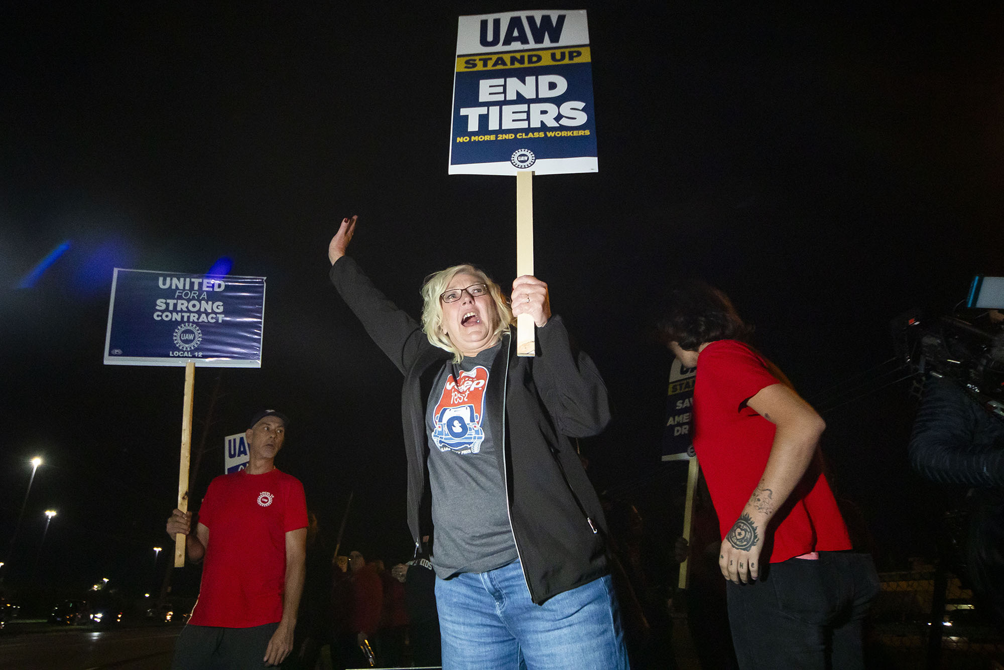 Photo: A white woman wearing a black jacket, tee, and jeans, shouts and raises a sign that reads "UAW Stand Up, End Tiers, No more second class workers".