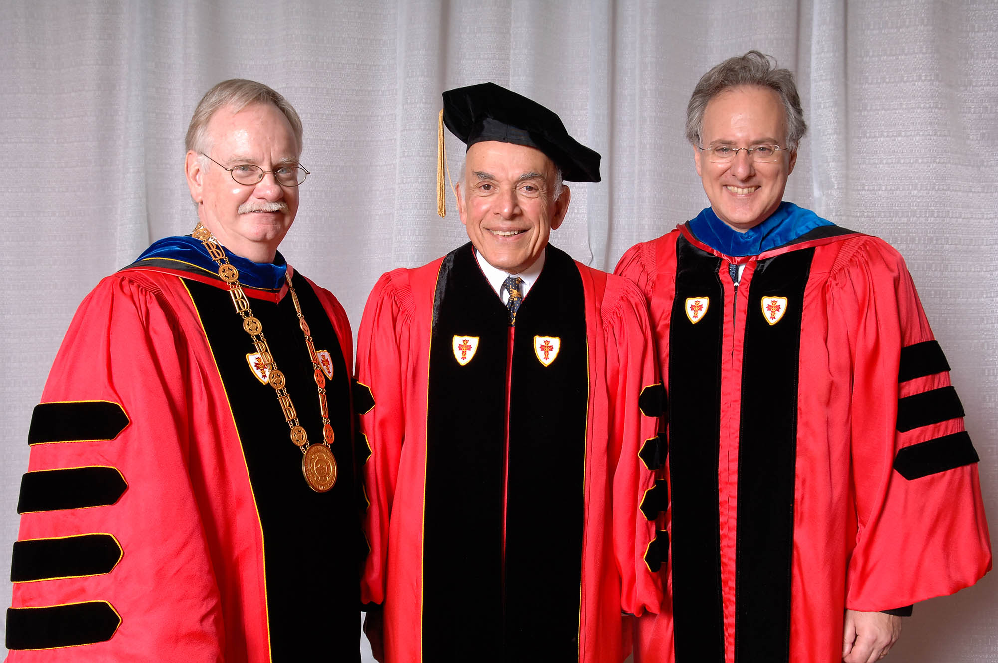 Photo: (From left to right) President Robert Brown, Aram Chobanian, and Trustee Alan Leventhal all wearing red and black ceremonial graduation gowns smile and pose in front of a grey backdrop. Brown wears a large gold medallion while Chobanian wears a black cap.