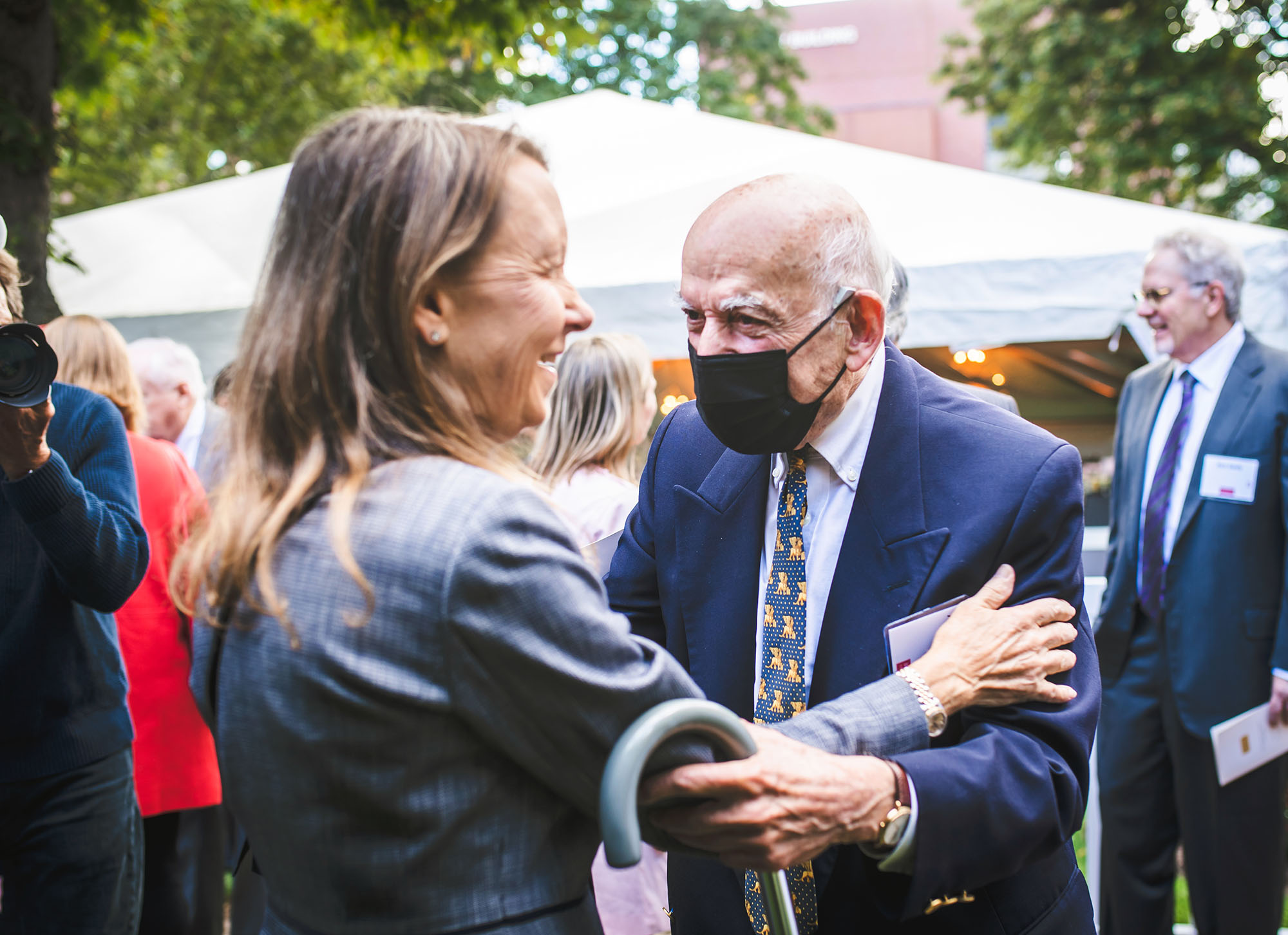 Photo: Aram Chobanian (right) greets Beverly Brown. An older man balding man with white hair and wearing a white collared shirt, blue and yellow tie, black face mask, and navy suit, looks cheerful as he holds a smiling brunette woman in grey suit.